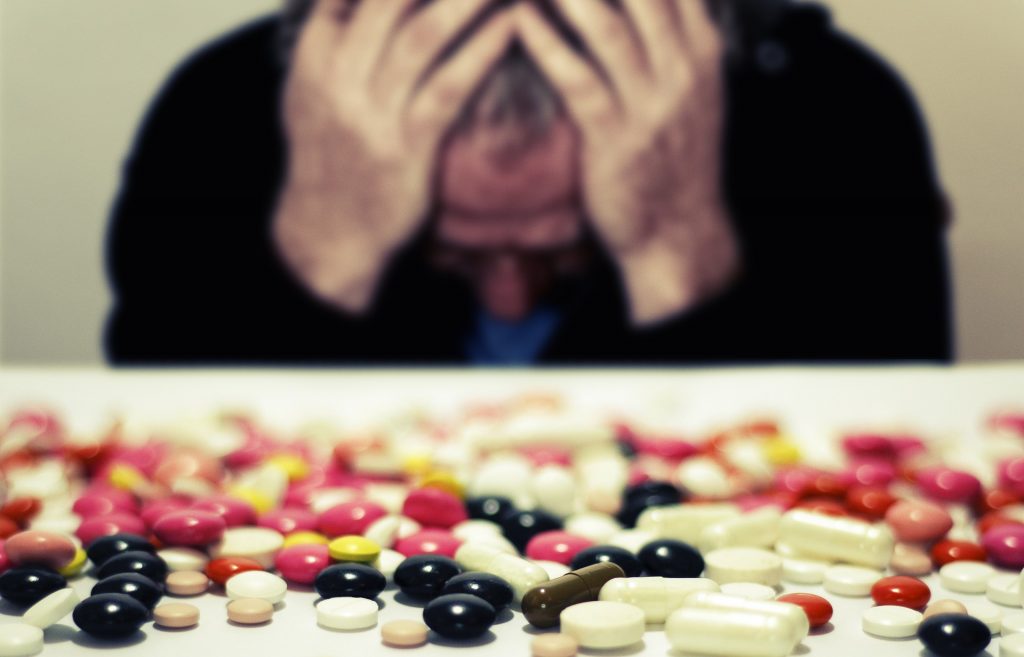 A distressed man holding his head in his hands with a pile of prescription drugs laid below him