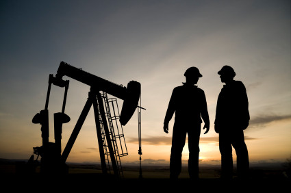 two workers stand talking in front of an oil field drill