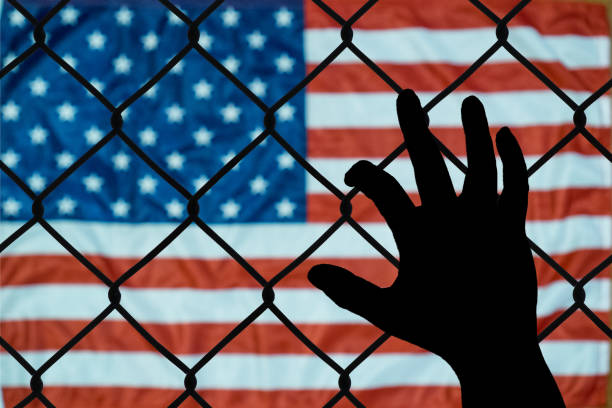 American flag and fence with a shadowed hand, representing U.S. immigration policy and border restrictions.